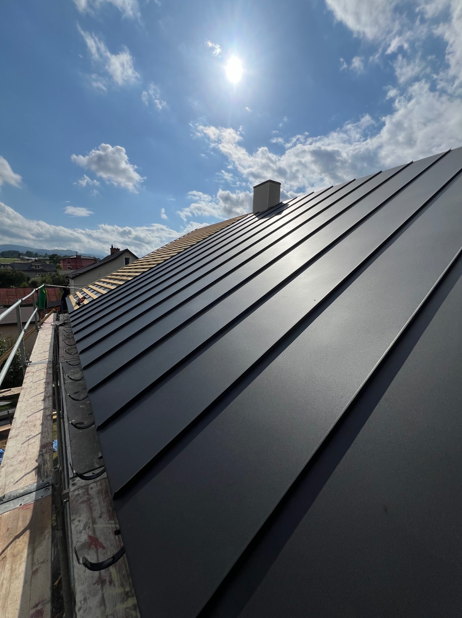 Nowy, grafitowy dach z blachy na tle błękitnego nieba z chmurami i fragmentem starego dachu. Widoczne rynny i komin. Słoneczny dzień.