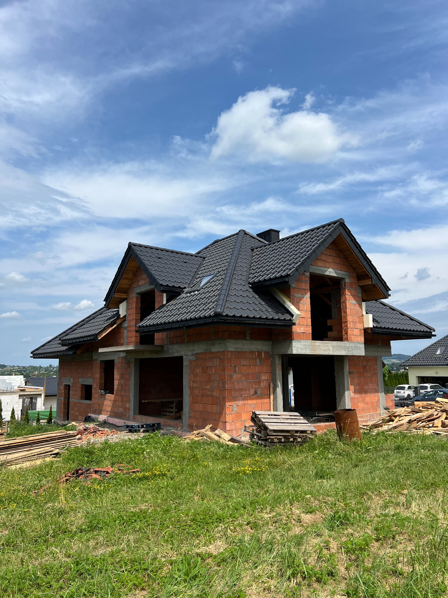Dom w trakcie budowy z czarną dachówką, widoczne cegły i betonowe elementy konstrukcyjne. W tle błękitne niebo z chmurami i zieleń trawy.