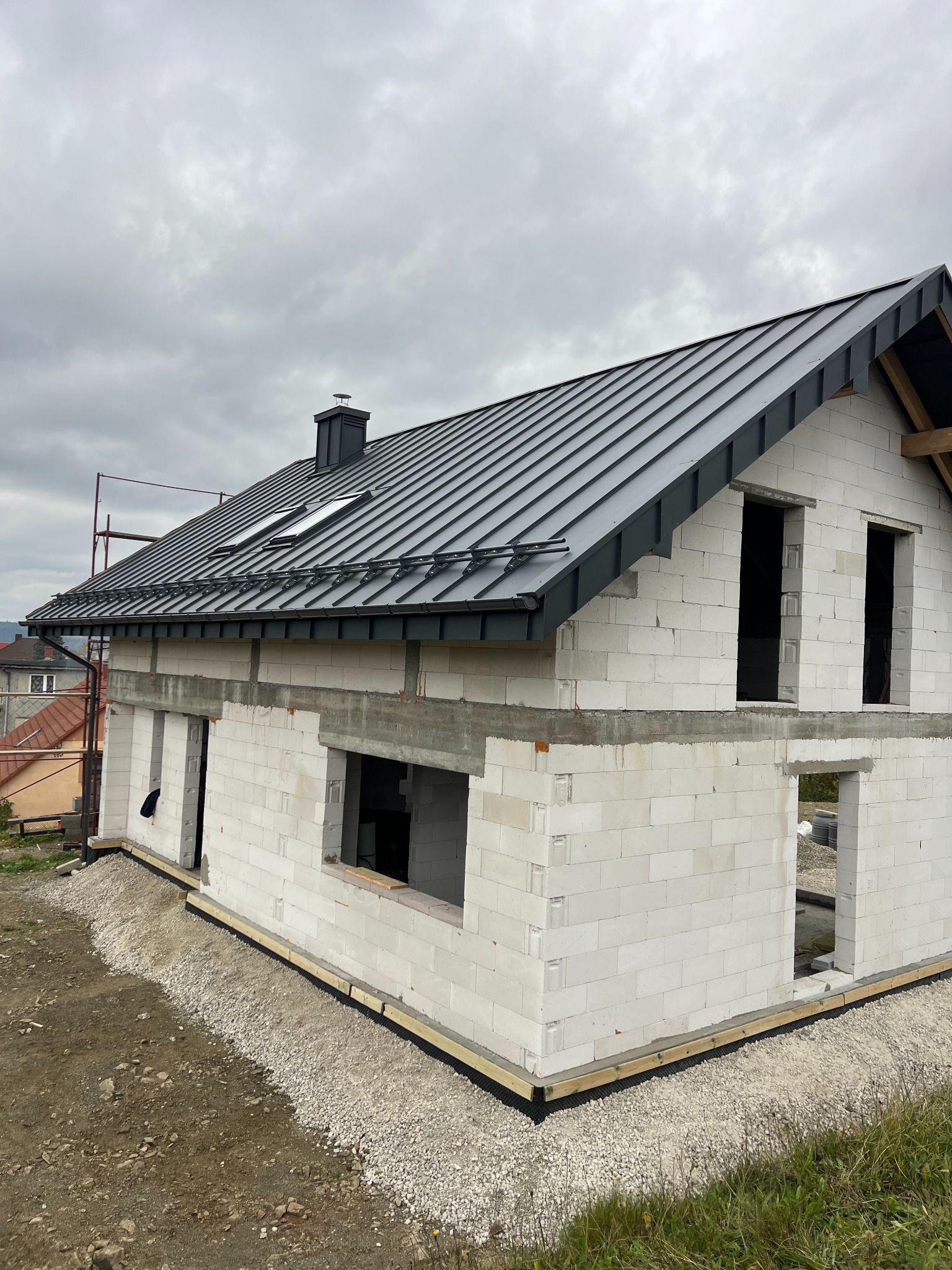 Surowy stan domu z białego bloczka z ciemnoszarym dachem z blachy na tle pochmurnego nieba. Widoczne okna dachowe i komin. Częściowo widoczne rusztowanie.