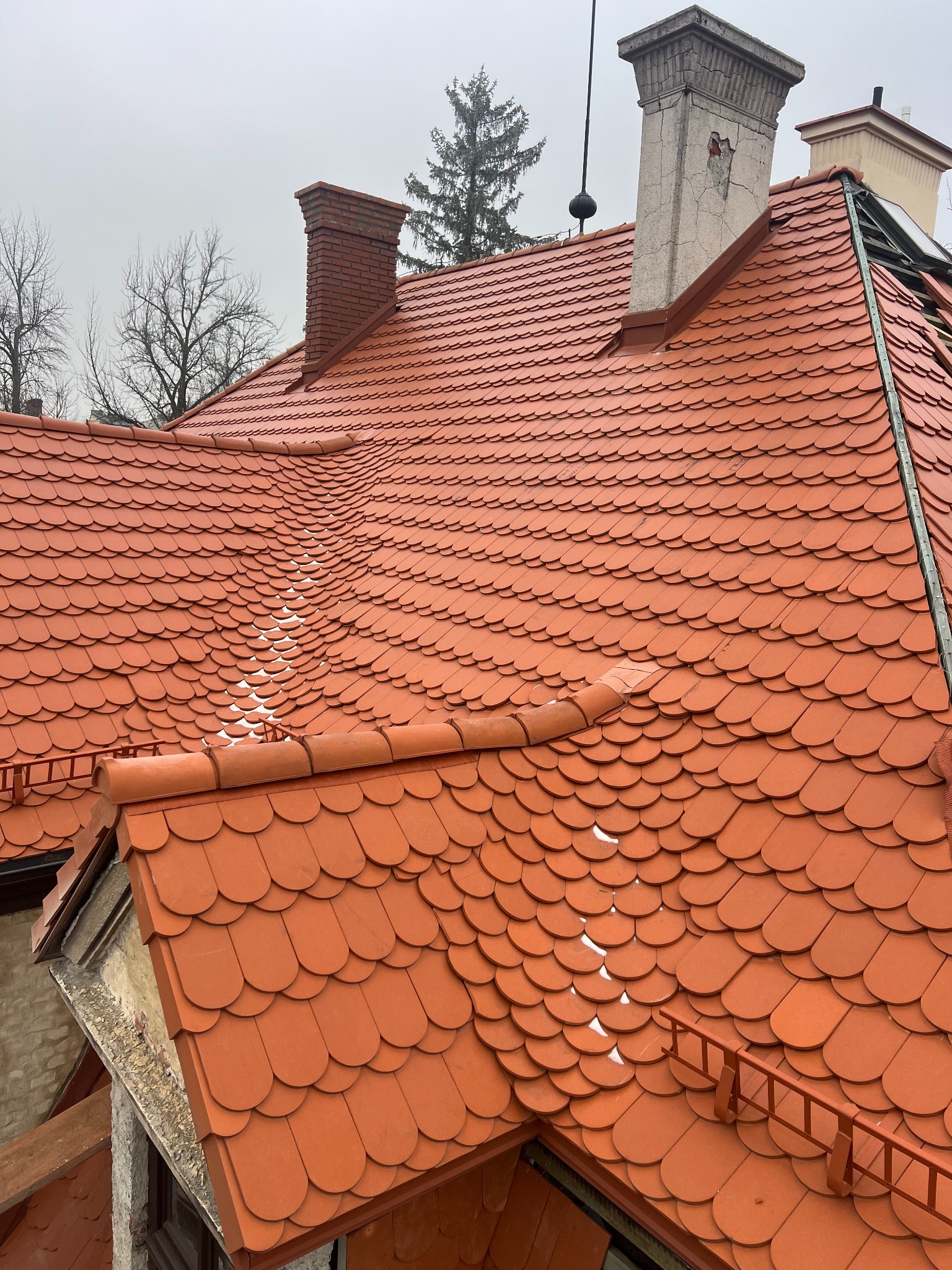 Czerwony dach z dachówki karpiówki, widoczne kominy i fragmenty instalacji odgromowej. Na dachu miejscami leży śnieg. Pochmurne niebo w tle.