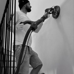 WALL in - Pracownik w czapce szlifuje ścianę szlifierką z odkurzaczem na schodach. Ujęcie od dołu, widoczne ślady farby na spodenkach.