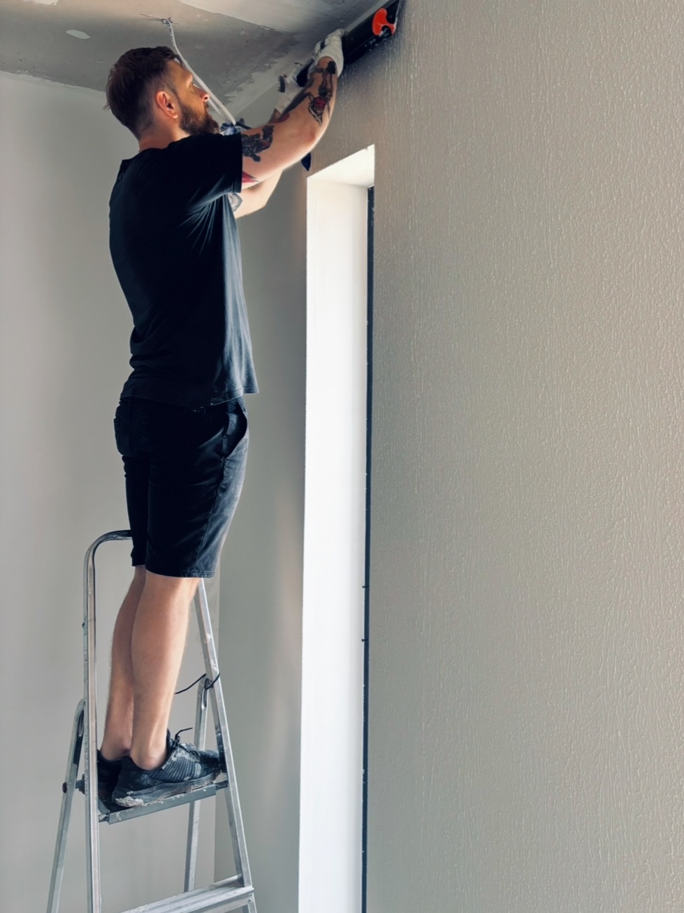 Mężczyzna na drabinie szpachluje ścianę szpachlą z pomarańczowym uchwytem. Widoczny fragment sufitu i ościeżnica okna. Na ramieniu tatuaż.