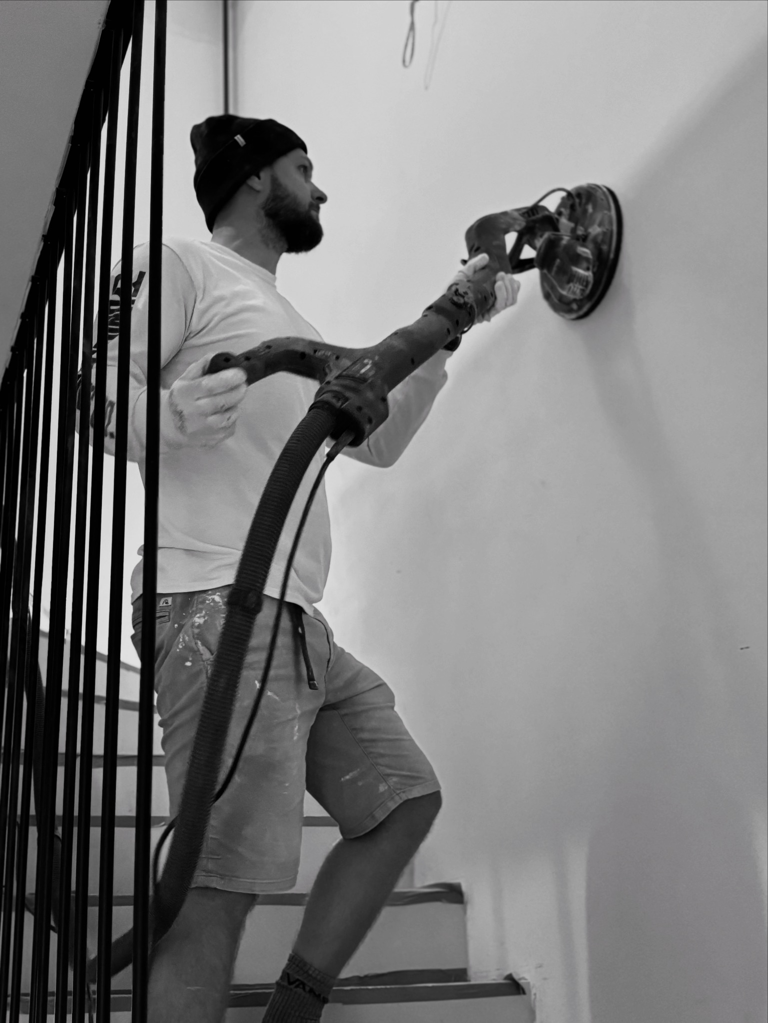 Pracownik w czapce szlifuje ścianę szlifierką z odkurzaczem na schodach. Ujęcie od dołu, widoczne ślady farby na spodenkach.