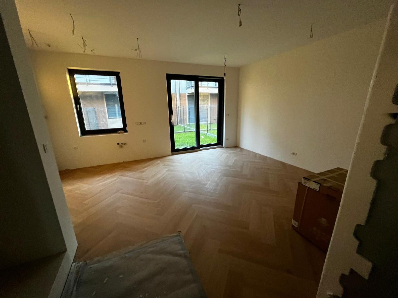 Jasne, parkietowe podłogi ułożone w jodełkę w pustym pokoju z oknami i drzwiami balkonowymi. Ściany przygotowane do malowania, widoczne kable oświetleniowe.