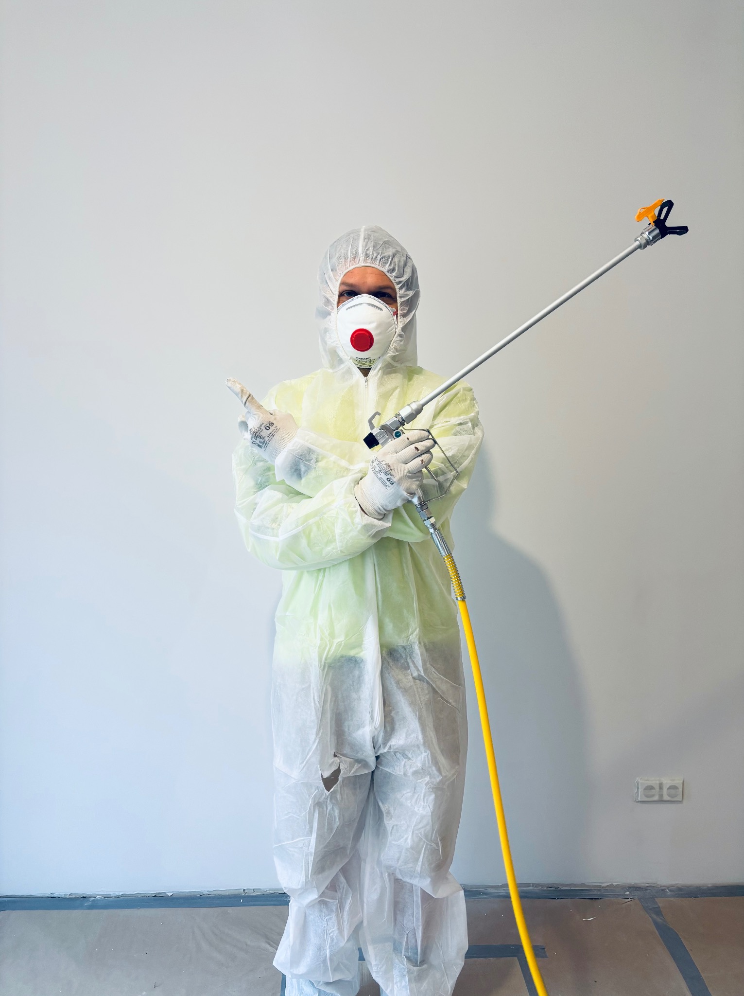Pracownik w kombinezonie ochronnym i masce trzymający pistolet do natrysku na tle białej ściany. Widoczny przewód i zabezpieczona podłoga.