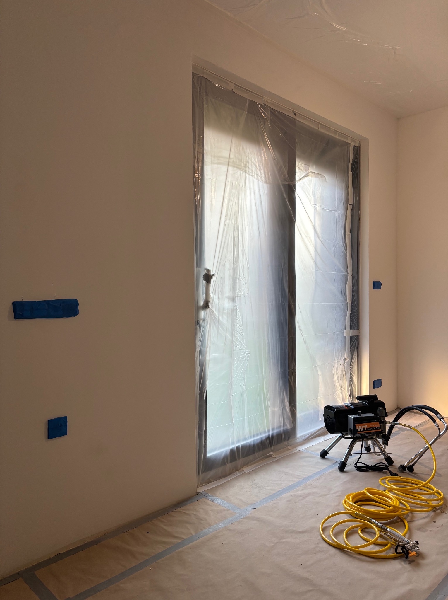 Przygotowane pomieszczenie do malowania natryskowego: okno zabezpieczone folią, ściany z taśmą maskującą, na podłodze papier ochronny, widoczny agregat malarski.