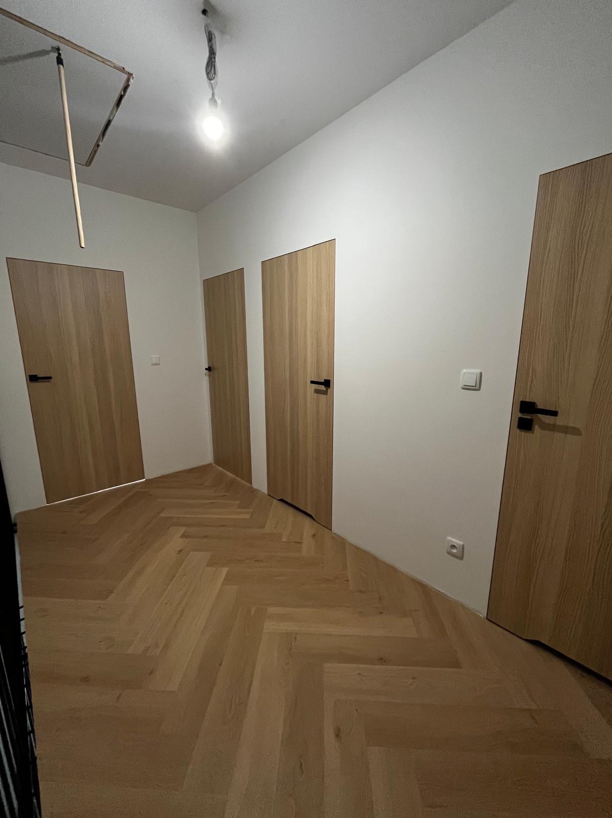 Jasne, odnowione wnętrze z parkietem w jodełkę, trzema drewnianymi drzwiami z czarnymi klamkami i świeżo pomalowanymi białymi ścianami. Widoczna instalacja oświetleniowa.