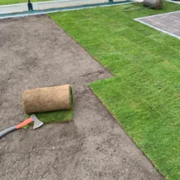 Rozkładanie trawy z rolki na przygotowanym gruncie. Widoczny fragment ułożonej trawy, rolka trawy oraz toporek. W tle ogrodzenie i budynek.