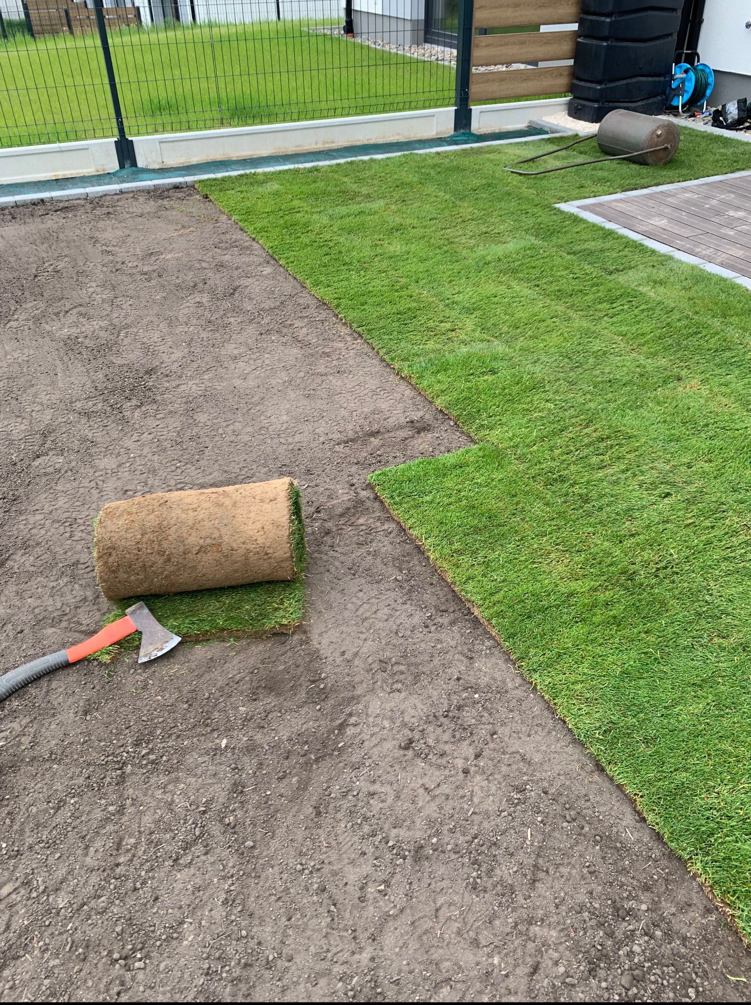 Rozkładanie trawy z rolki na przygotowanym gruncie. Widoczny fragment ułożonej trawy, rolka trawy oraz toporek. W tle ogrodzenie i budynek.