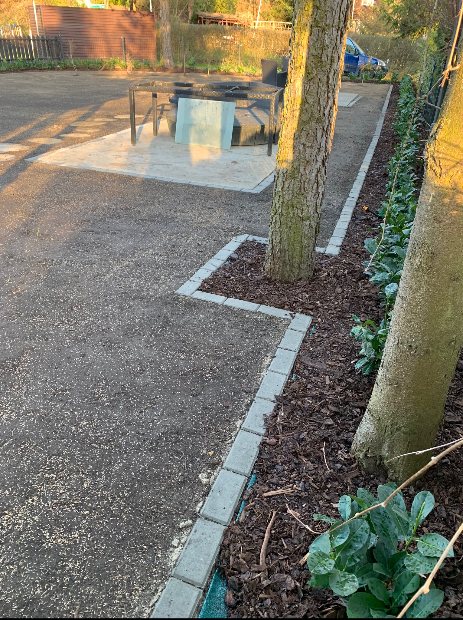 Fragment świeżo założonego ogrodu z obrzeżami z kostki, korą wokół drzew i nowo posianą trawą. W tle meble ogrodowe i fragment żywopłotu.