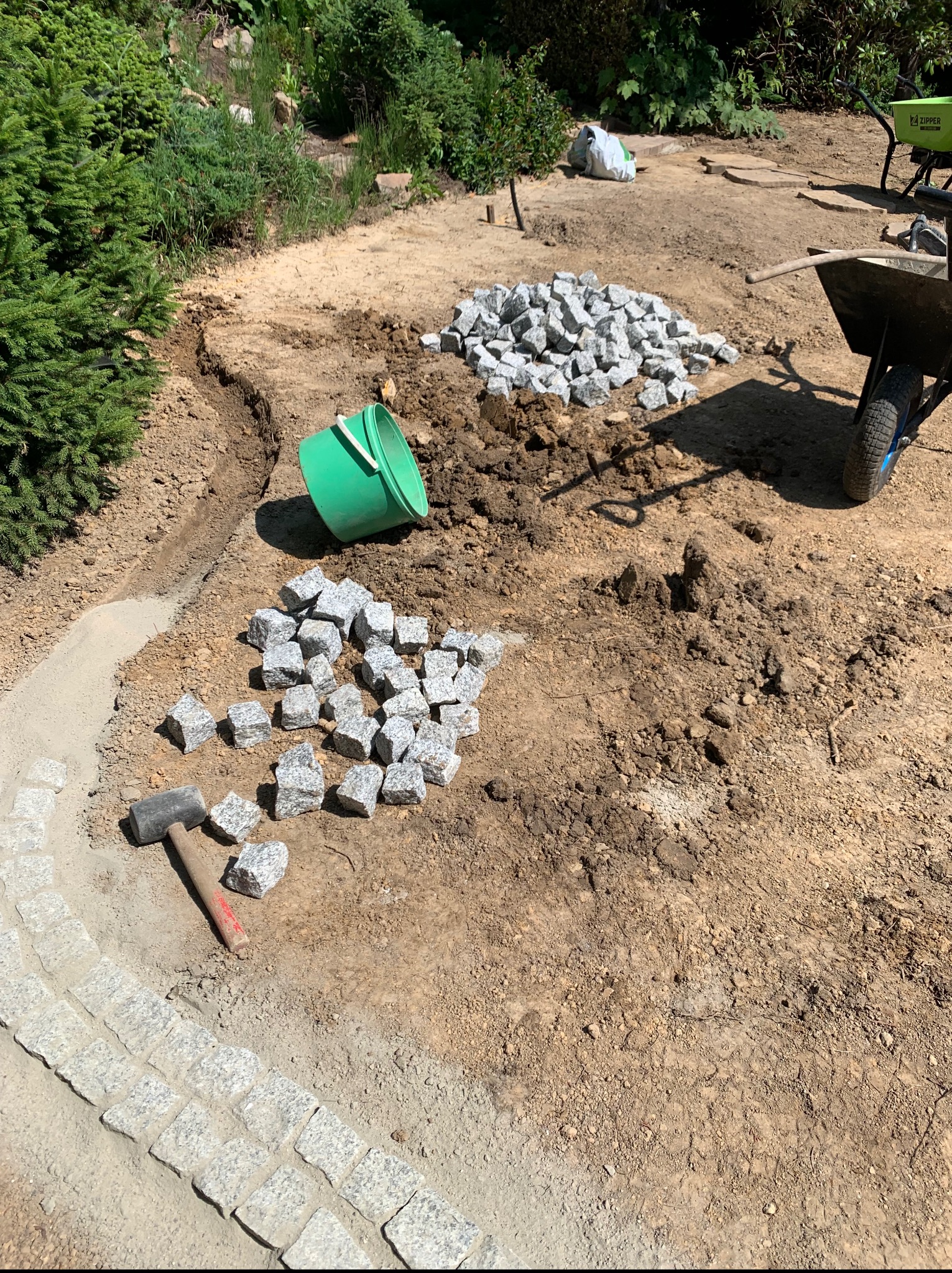 Układanie kostki granitowej na ścieżce w ogrodzie. Widoczne narzędzia: gumowy młotek, wiadro i taczka. Częściowo ułożona ścieżka z kostki.