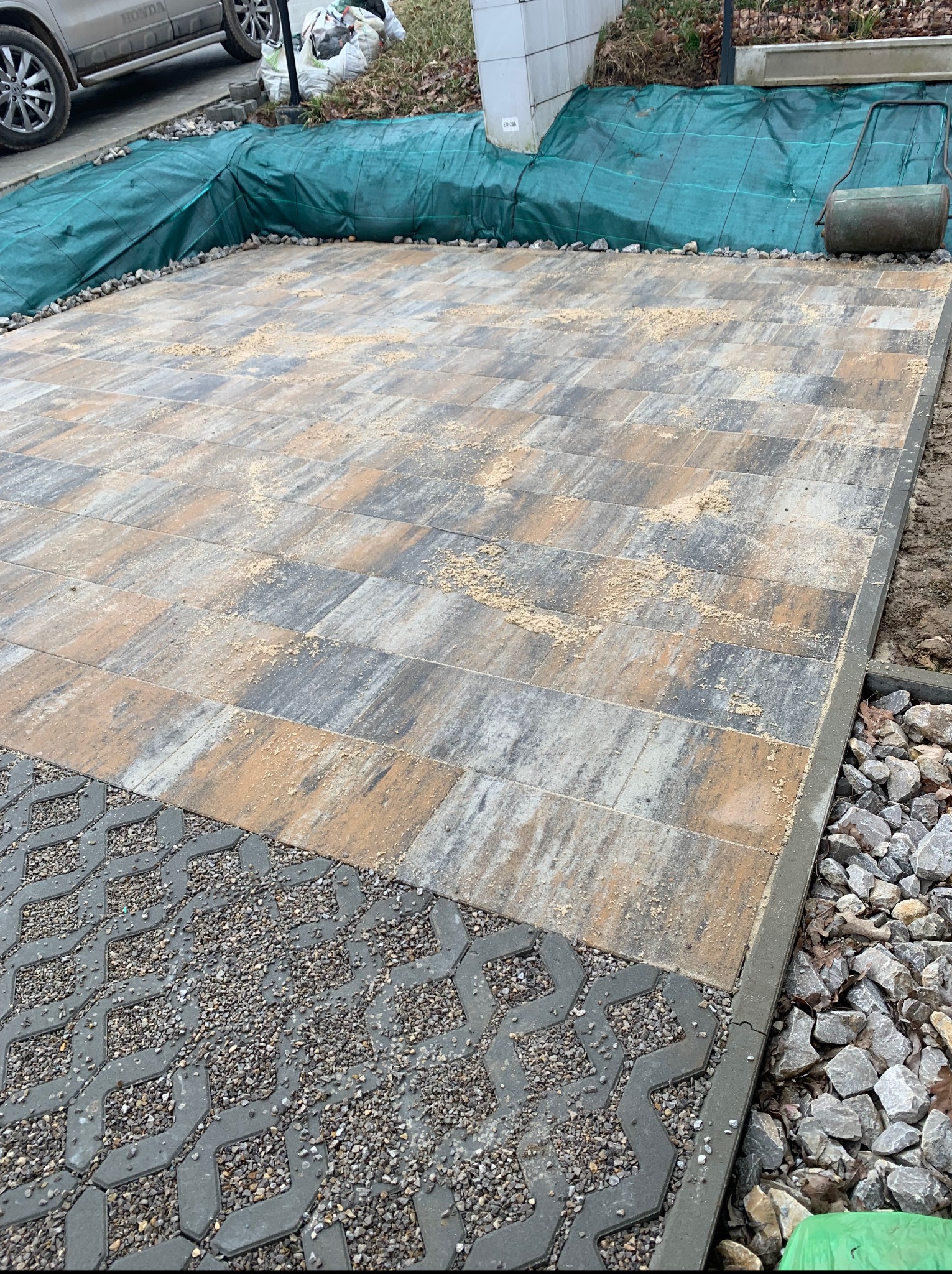 Układanie kostki brukowej na posesji, widoczne obrzeża i podsypka piaskowa. Część powierzchni wyłożona ażurowymi płytami wypełnionymi grysem.