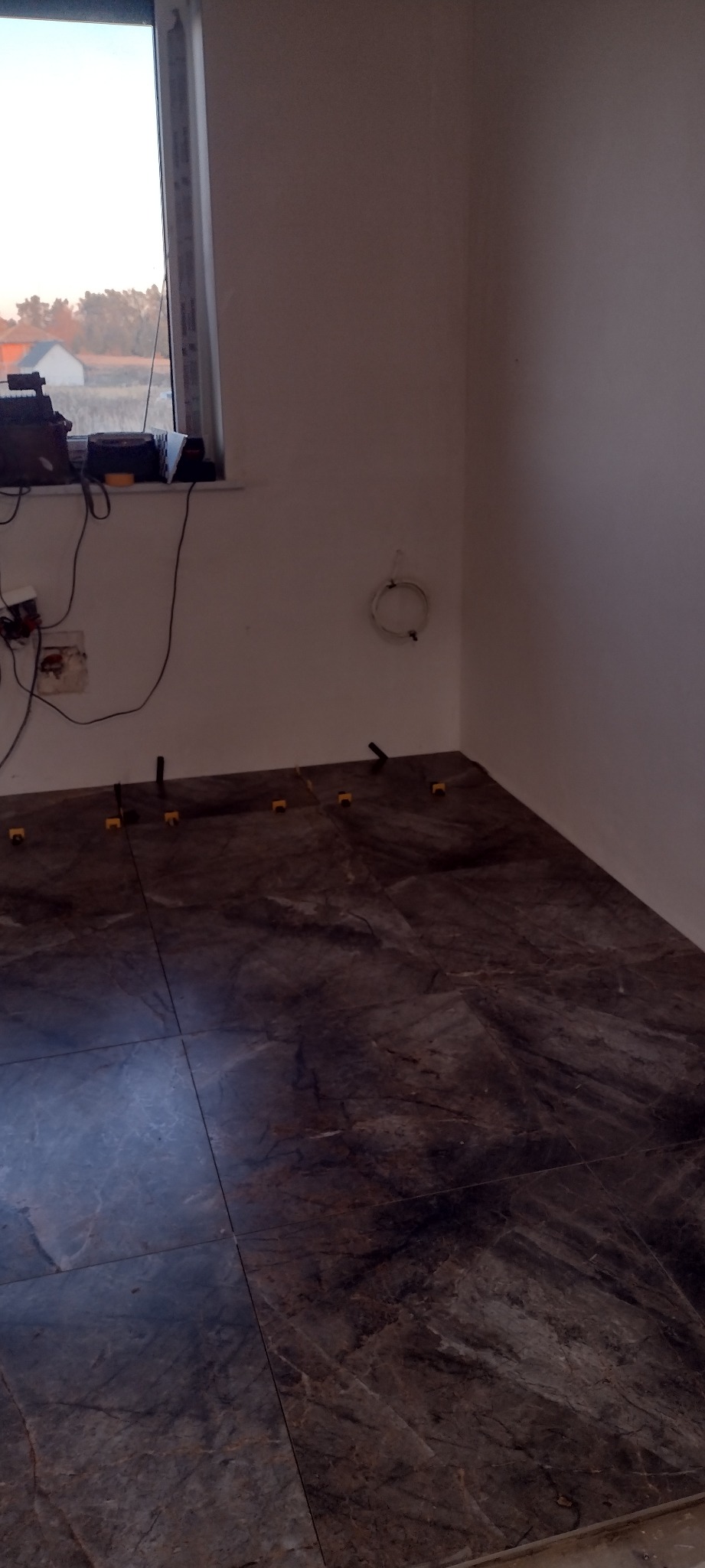 Ułożone płytki podłogowe imitujące marmur, z żółtymi krzyżykami dystansowymi, w rogu pokoju z oknem. Widoczne narzędzia i kable na parapecie.