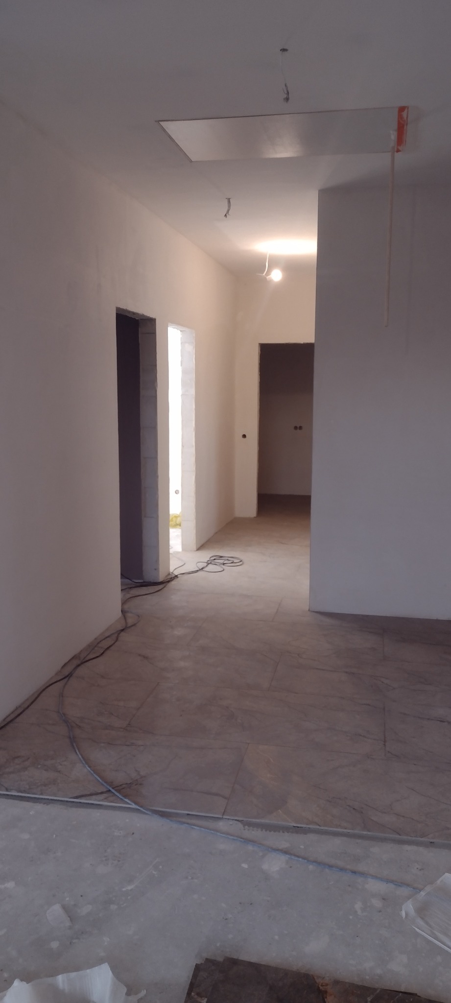 Wnętrze w trakcie prac wykończeniowych: świeżo pomalowane ściany na biało, ułożona podłoga z płytek imitujących marmur, widoczne kable i otwory drzwiowe.