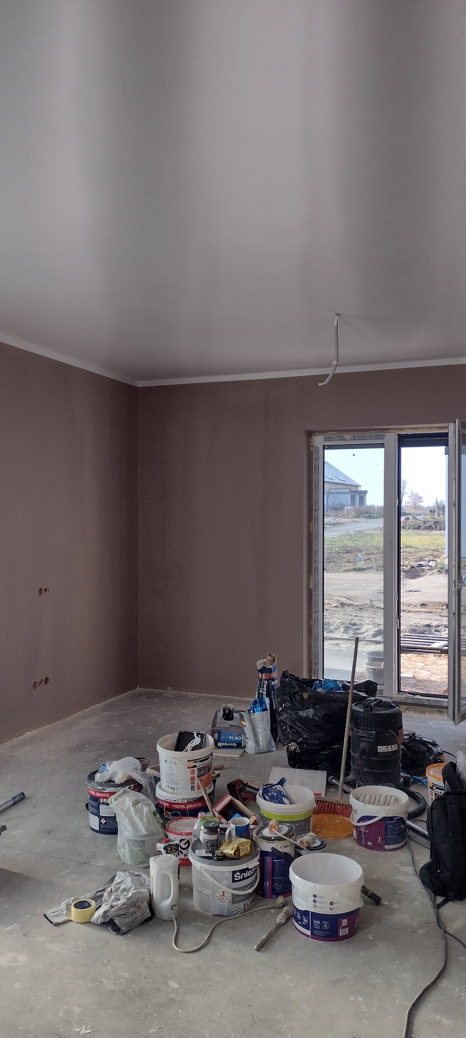 Pokój po gładzi, przygotowany do malowania. Widoczne puszki z farbą, narzędzia malarskie i worki na podłodze. Ściany w kolorze beżowym, sufit biały. Okno balkonowe z widokiem na zewnątrz.