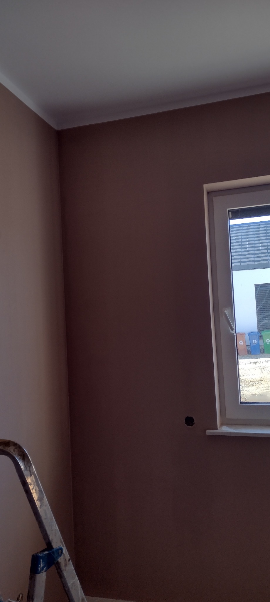 Ściana w odcieniach brązu po malowaniu, widoczne białe listwy przysufitowe i fragment drabiny malarskiej. W tle okno z widokiem na zewnątrz.