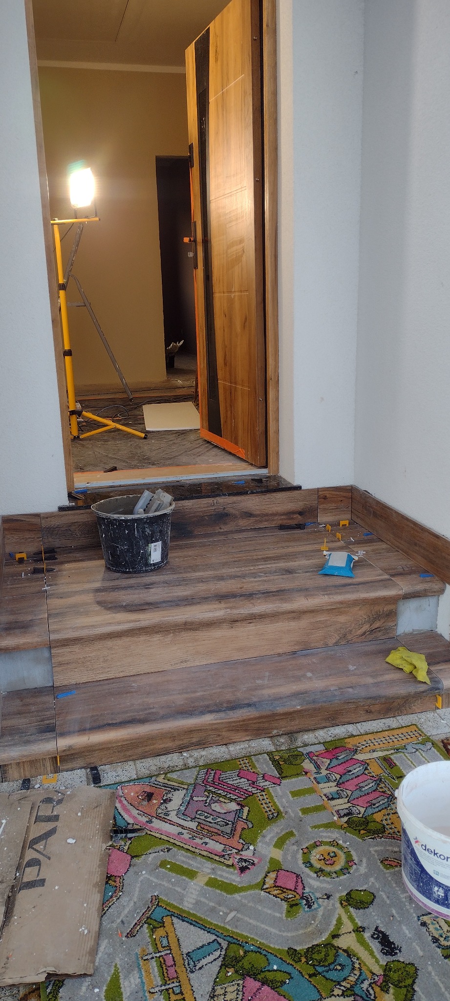 Układanie płytek imitujących drewno na schodach wejściowych. Widoczny kubeł z zaprawą, poziomica i fragment dziecięcego dywanu. Oświetlenie halogenowe w tle.