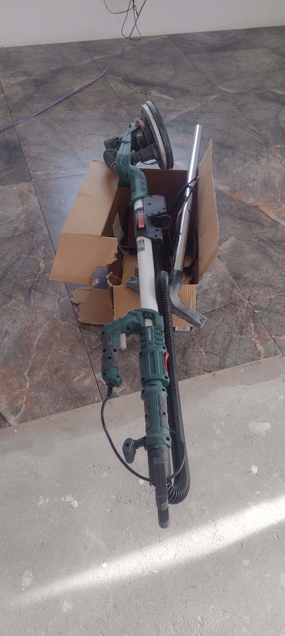 Szlifierka do gipsu Parkside w kartonie, z widocznym odkurzaczem i przewodem. Sprzęt na podłodze z płytek i surowego betonu. Widok z góry.