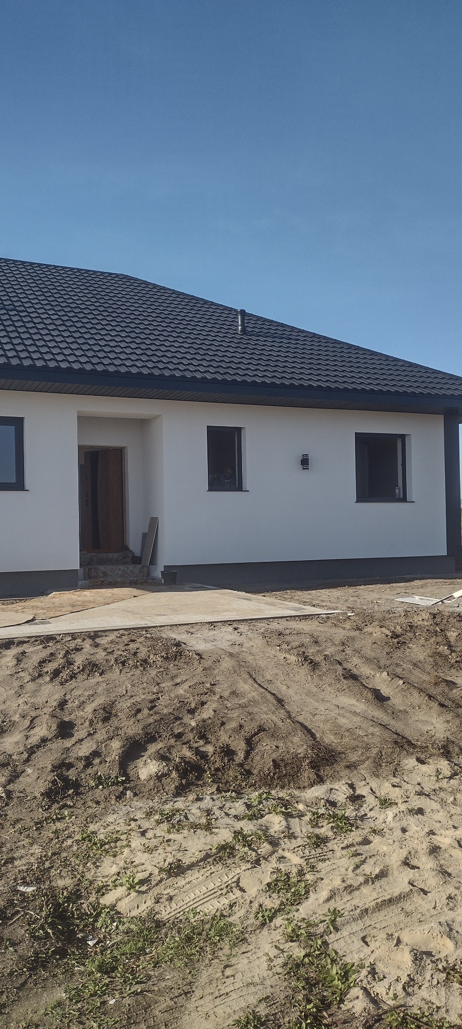 Biały dom z ciemną dachówką i oknami, widok z zewnątrz, teren wokół domu w trakcie prac ziemnych, błękitne niebo w tle. Minimalistyczny styl.