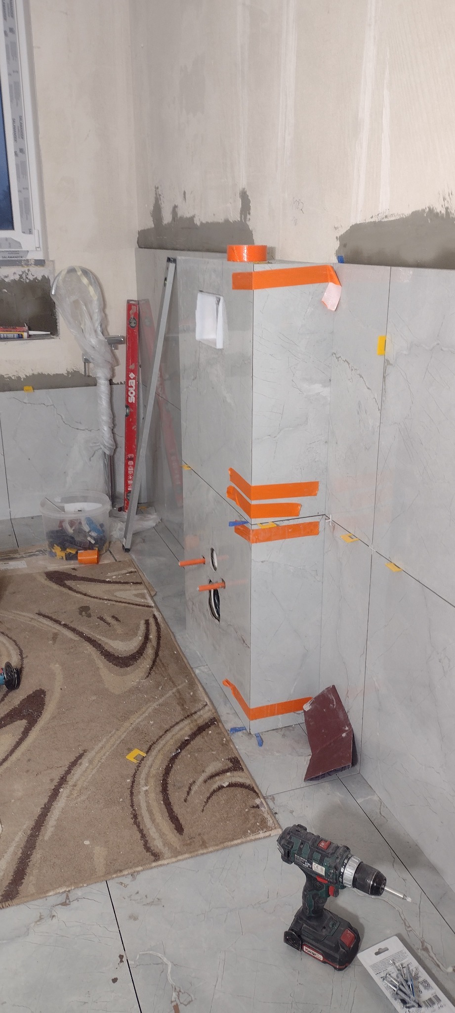 Montaż płytek imitujących marmur w łazience, zabezpieczonych pomarańczową taśmą. Widoczne narzędzia: poziomica, wiertarka. Fragment dywanu na podłodze.