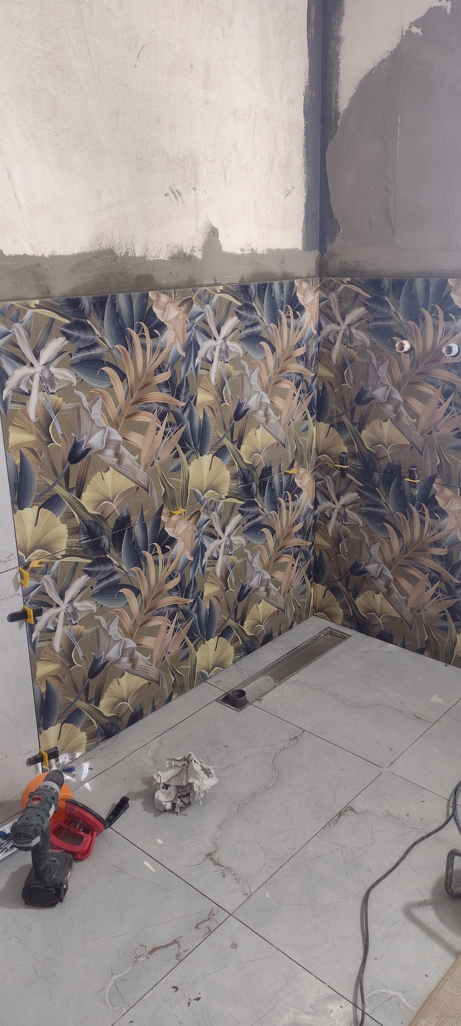 Układanie płytek z motywem roślinnym w łazience. Widoczny odpływ liniowy, narzędzia glazurnicze i fragment surowej ściany. Prace w toku.