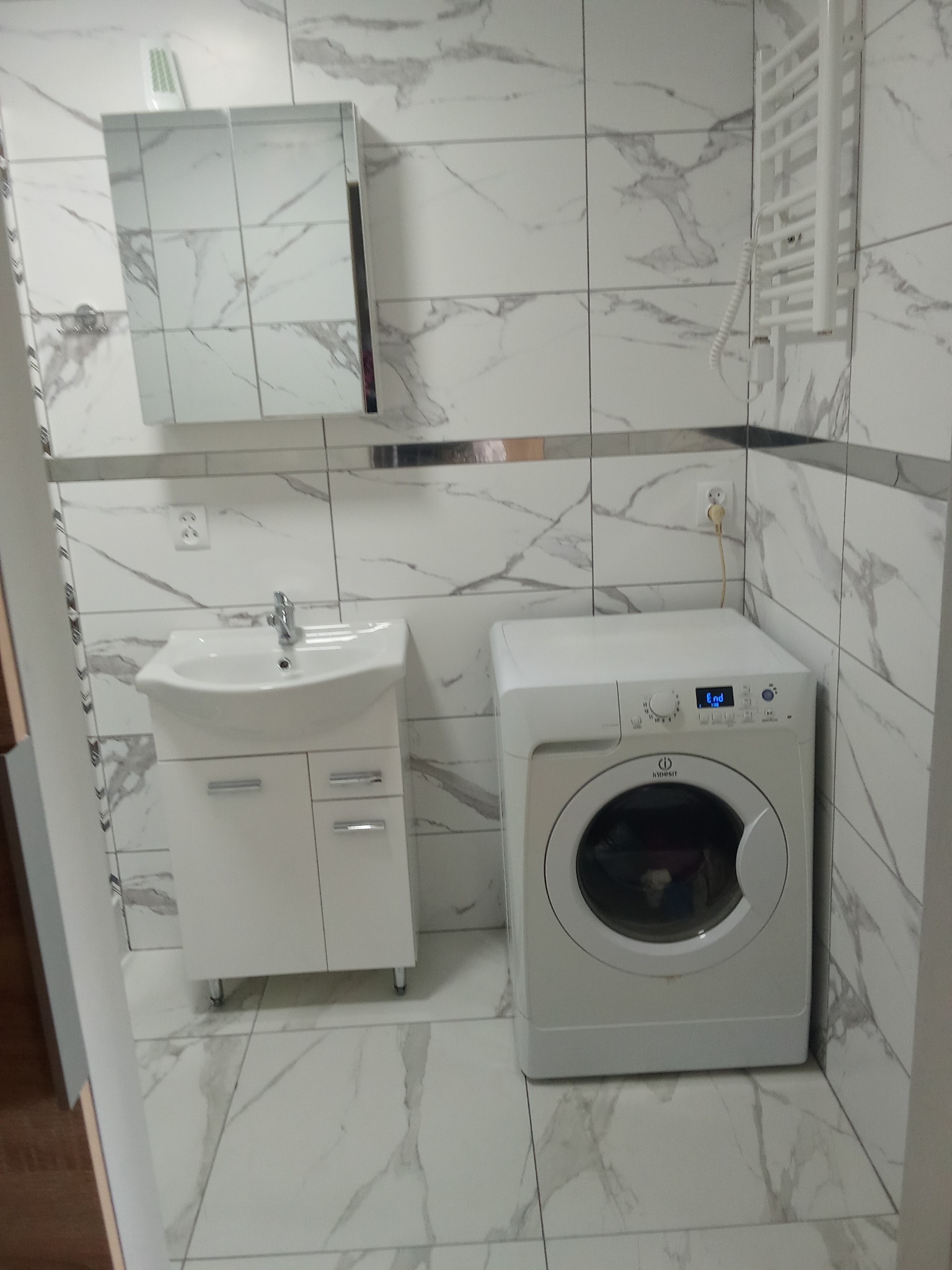 Biała łazienka z marmurowymi płytkami: umywalka z szafką, pralka i grzejnik drabinkowy. Minimalistyczny design z srebrnymi akcentami.