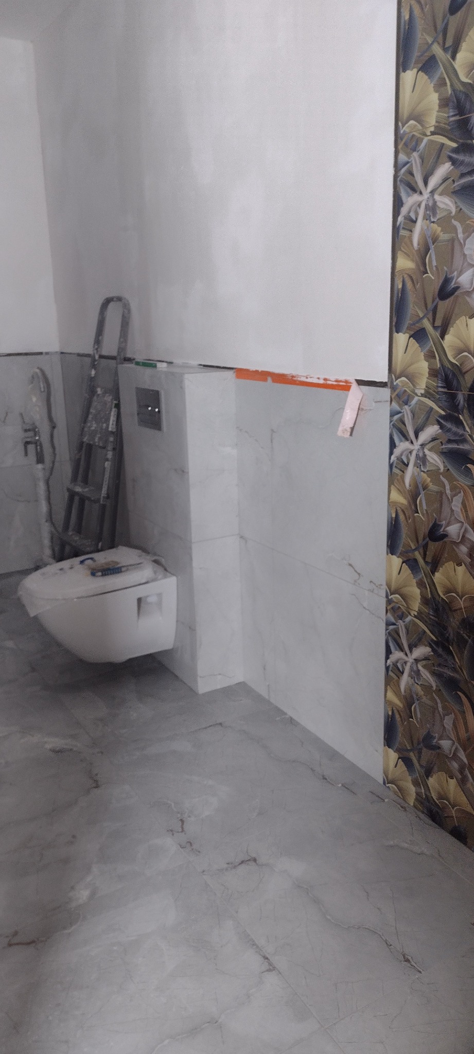 Montaż podwieszanej toalety w łazience z marmurowymi płytkami i dekoracyjną ścianą. Widoczna drabina i narzędzia, co sugeruje trwający remont.