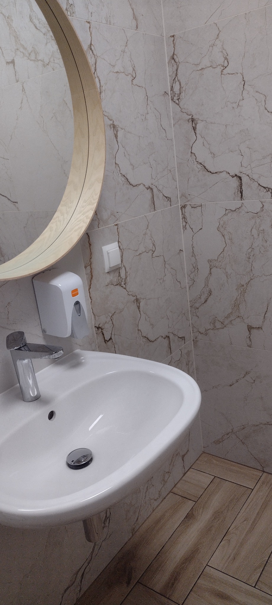 Nowoczesna łazienka z umywalką, okrągłym lustrem w drewnianej ramie i marmurowymi płytkami. Widoczny dozownik mydła i chromowana bateria.