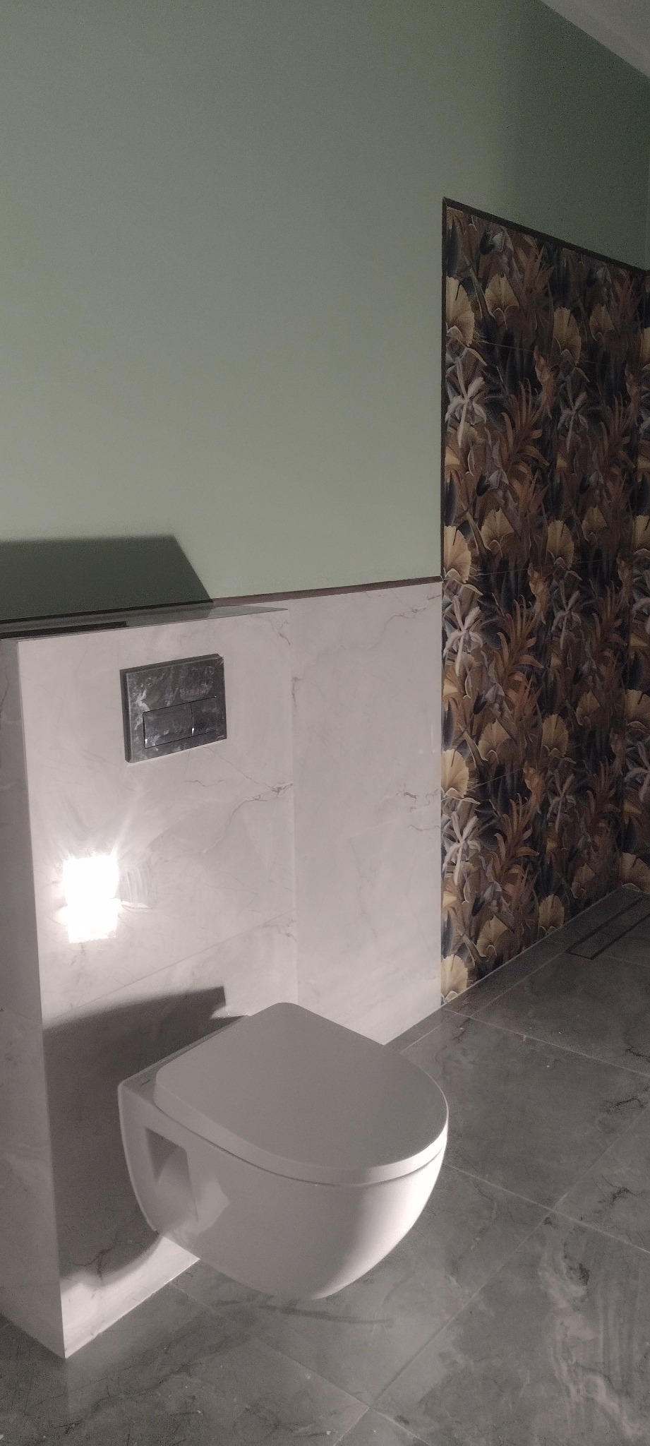 Nowoczesna toaleta podwieszana w wykończonej łazience. Marmurowe płytki ścienne i podłogowe, ściana akcentowa z tapetą w tropikalne liście. Minimalistyczny design.