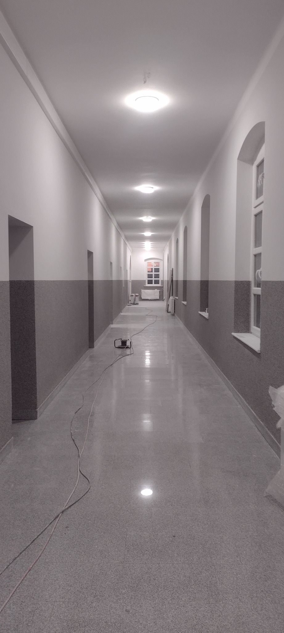 Długi korytarz po remoncie z dwukolorowymi ścianami, oświetleniem sufitowym i sprzętem budowlanym. Widok w perspektywie, podkreślający przestrzeń i świeżość wykończenia.