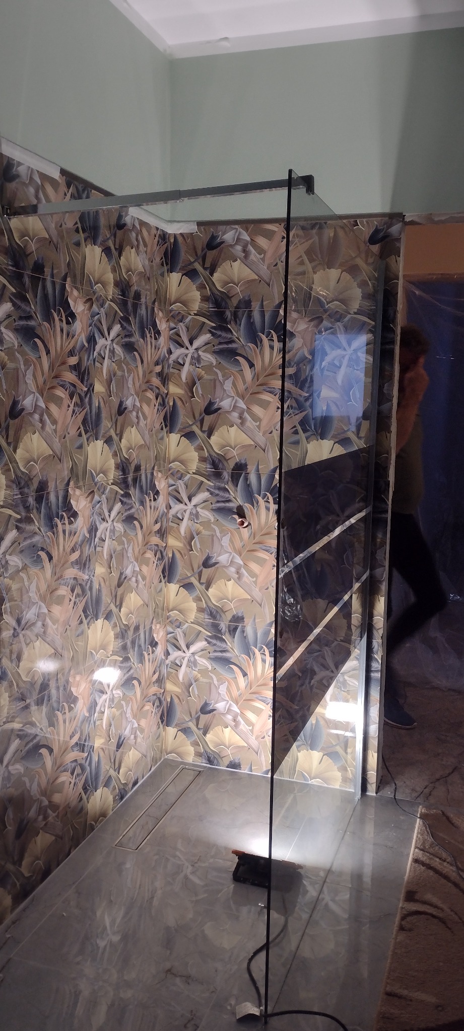 Wykończona kabina prysznicowa z wzorzystymi płytkami w odcieniach beżu i granatu. Widoczny odpływ liniowy w posadzce i częściowo widoczna osoba w tle.