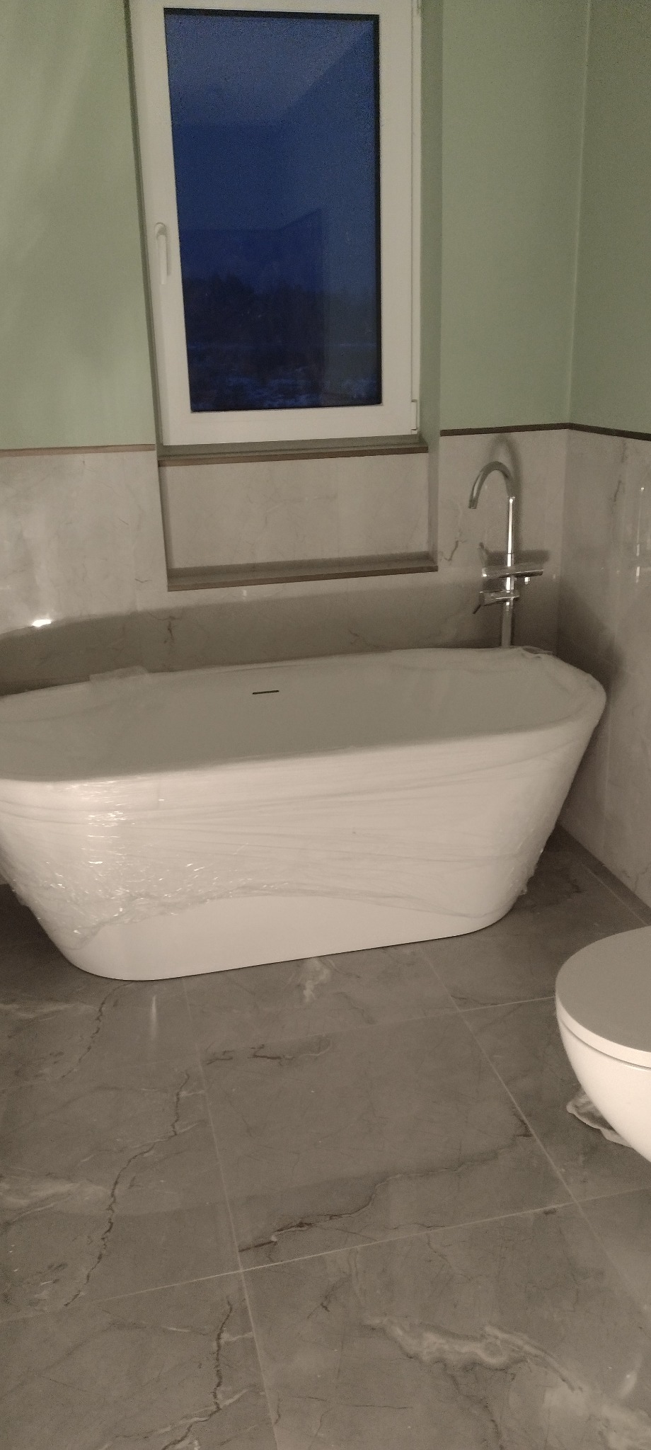 Nowoczesna, biała wanna owalna, zabezpieczona folią, w łazience z marmurowymi płytkami i oknem. Widoczna bateria i fragment toalety. Stonowana kolorystyka.