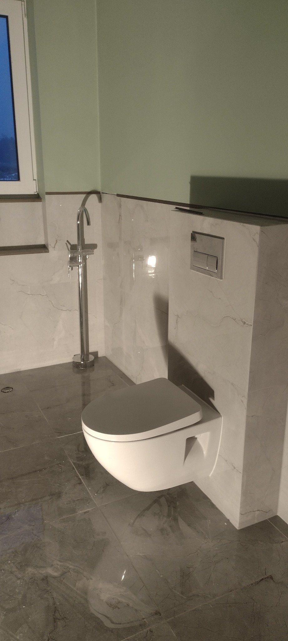 Nowoczesna, podwieszana toaleta z chromowaną armaturą na tle marmurowych płytek w odcieniach szarości i bieli. Minimalistyczny design, eleganckie wykończenie łazienki.