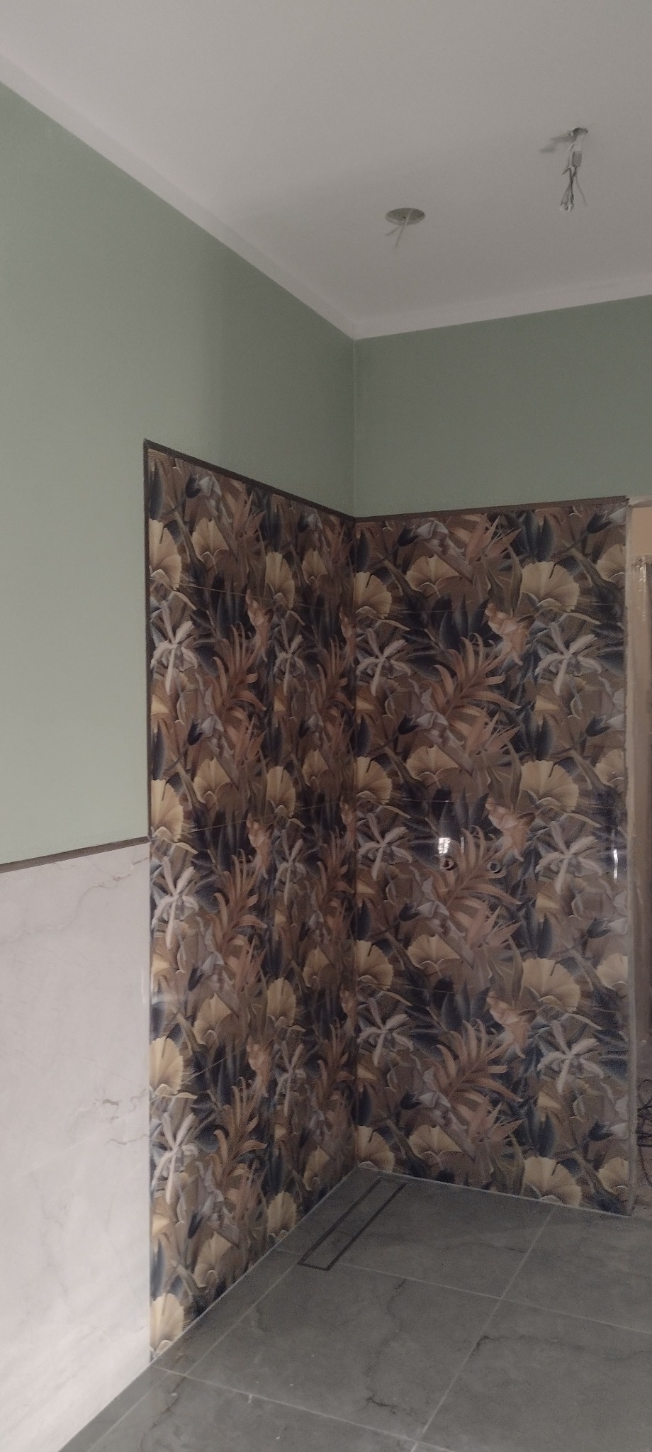 Wykończona łazienka z płytkami w kwiatowy wzór na ścianach i szarą podłogą. Widoczne fragmenty zielonej ściany i białej listwy. Oświetlenie sufitowe.