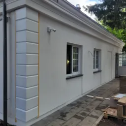 Otynkowany budynek z dekoracyjnymi elementami architektonicznymi, świeżo po malowaniu. Widoczne okna, drzwi i fragment chodnika z kostki.