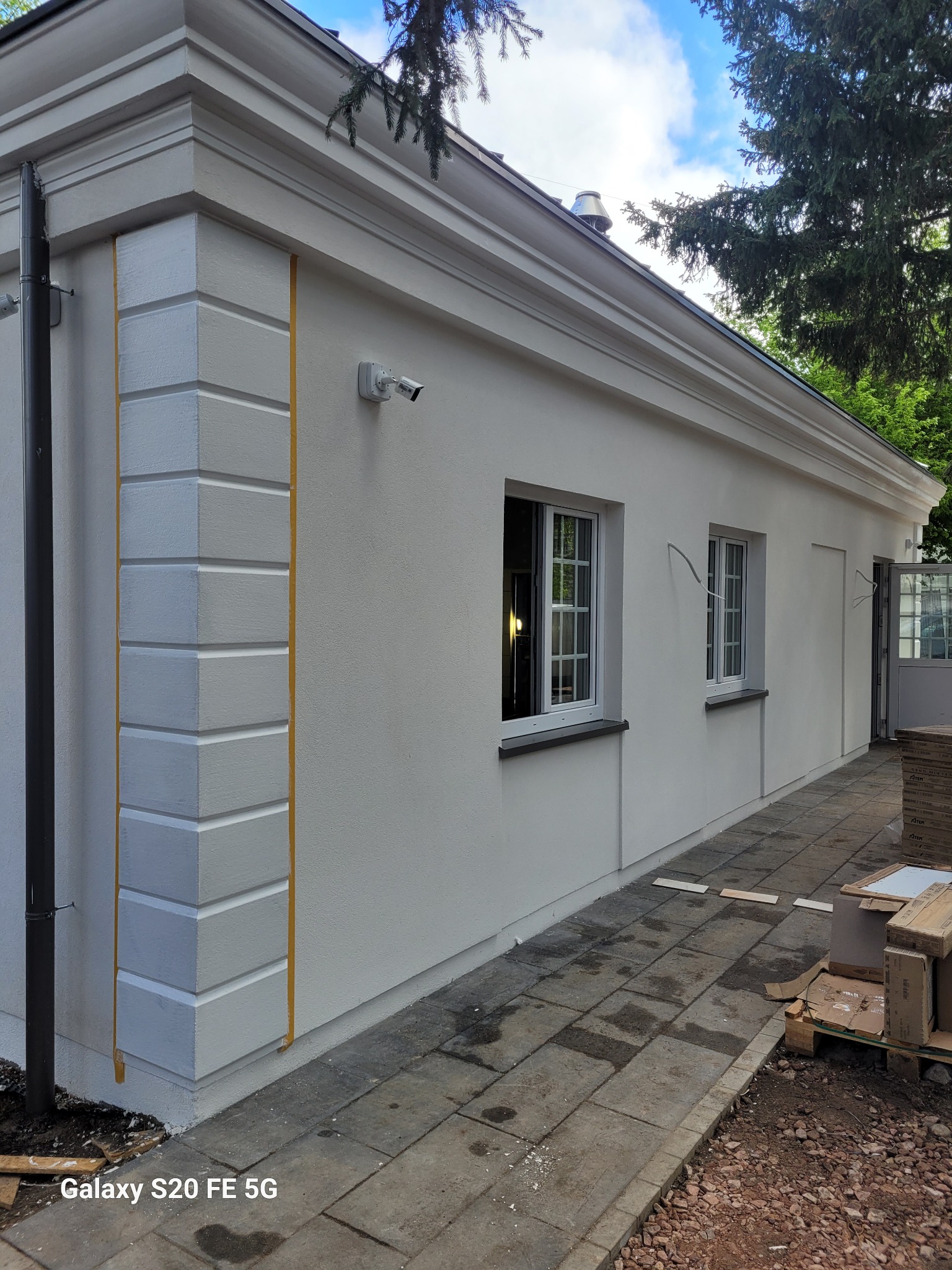 Otynkowany budynek z dekoracyjnymi elementami architektonicznymi, świeżo po malowaniu. Widoczne okna, drzwi i fragment chodnika z kostki.