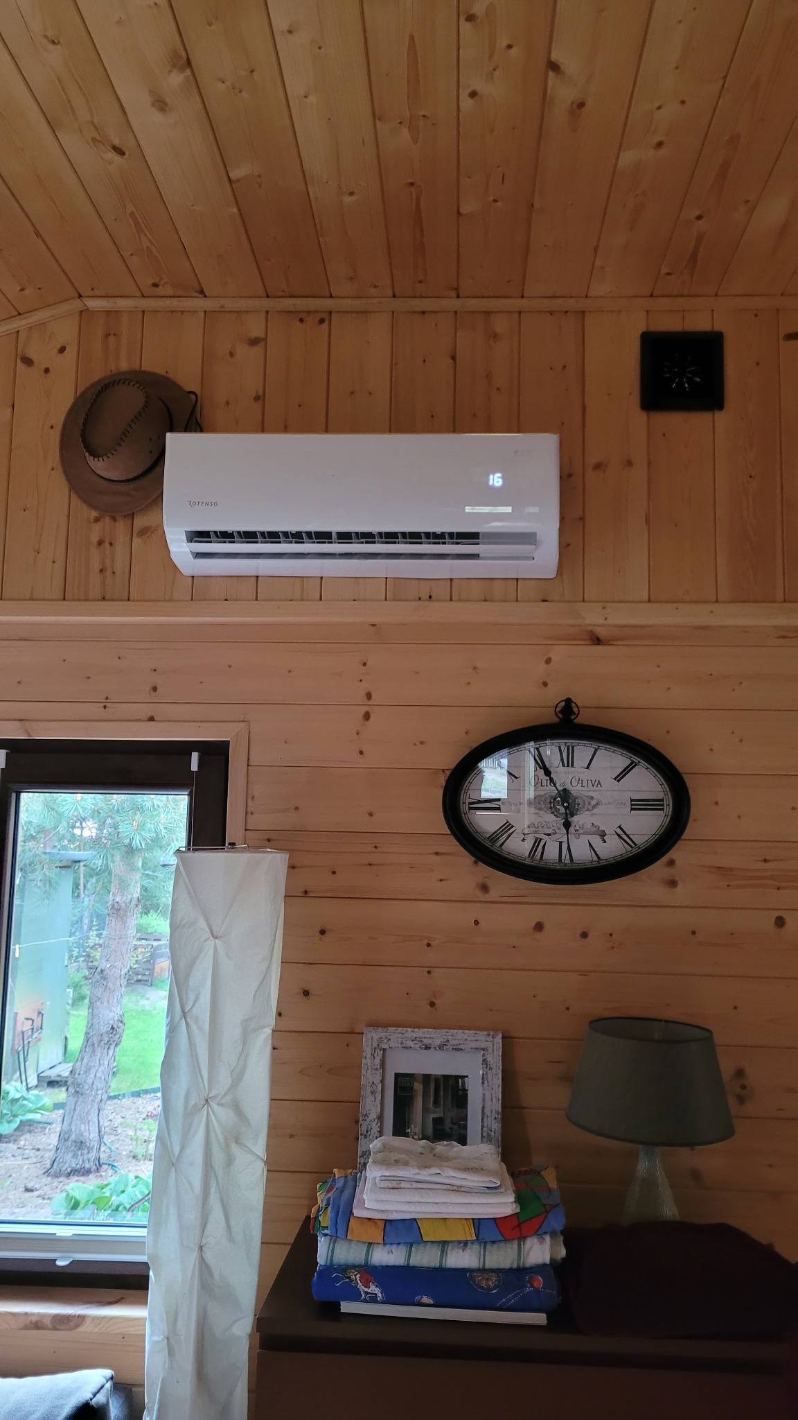 Klimatyzator Rotenso zamontowany na drewnianej ścianie w jasnym wnętrzu, obok wiszący kapelusz i zegar. Widoczny fragment okna z widokiem na zewnątrz.