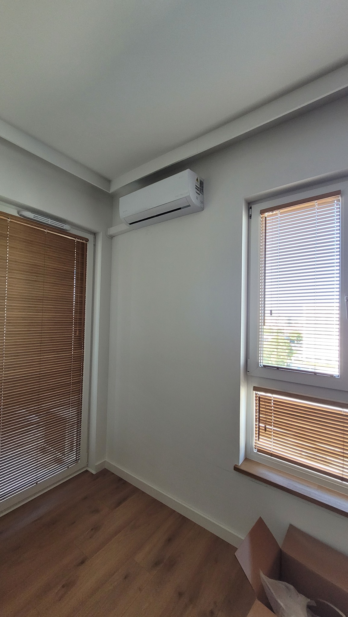 Biała klimatyzacja na ścianie w jasnym pokoju z drewnianą podłogą i żaluzjami w oknach. Kartonowe pudło w rogu pokoju sugeruje niedawny montaż.