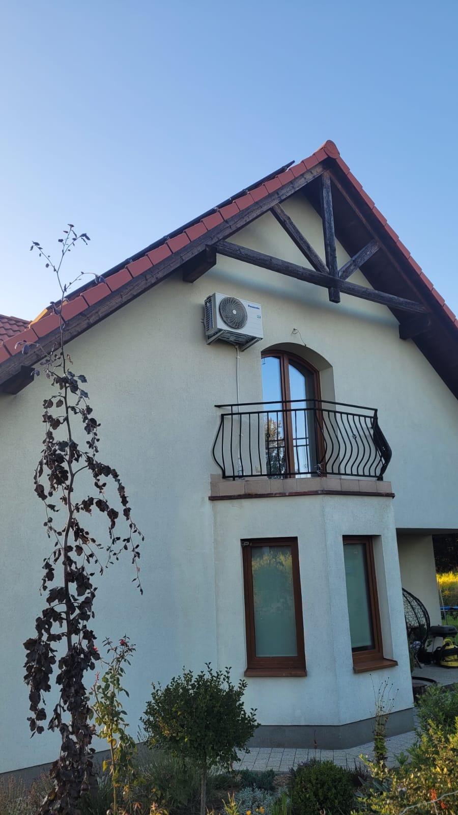 Biały klimatyzator Panasonic zamontowany na ścianie domu z balkonem. Widok z dołu, na tle błękitnego nieba.