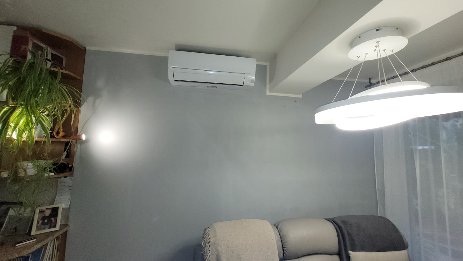 Biały klimatyzator na szarej ścianie w salonie. Widoczny nowoczesny żyrandol i fragment sofy. Roślina na półce po lewej.