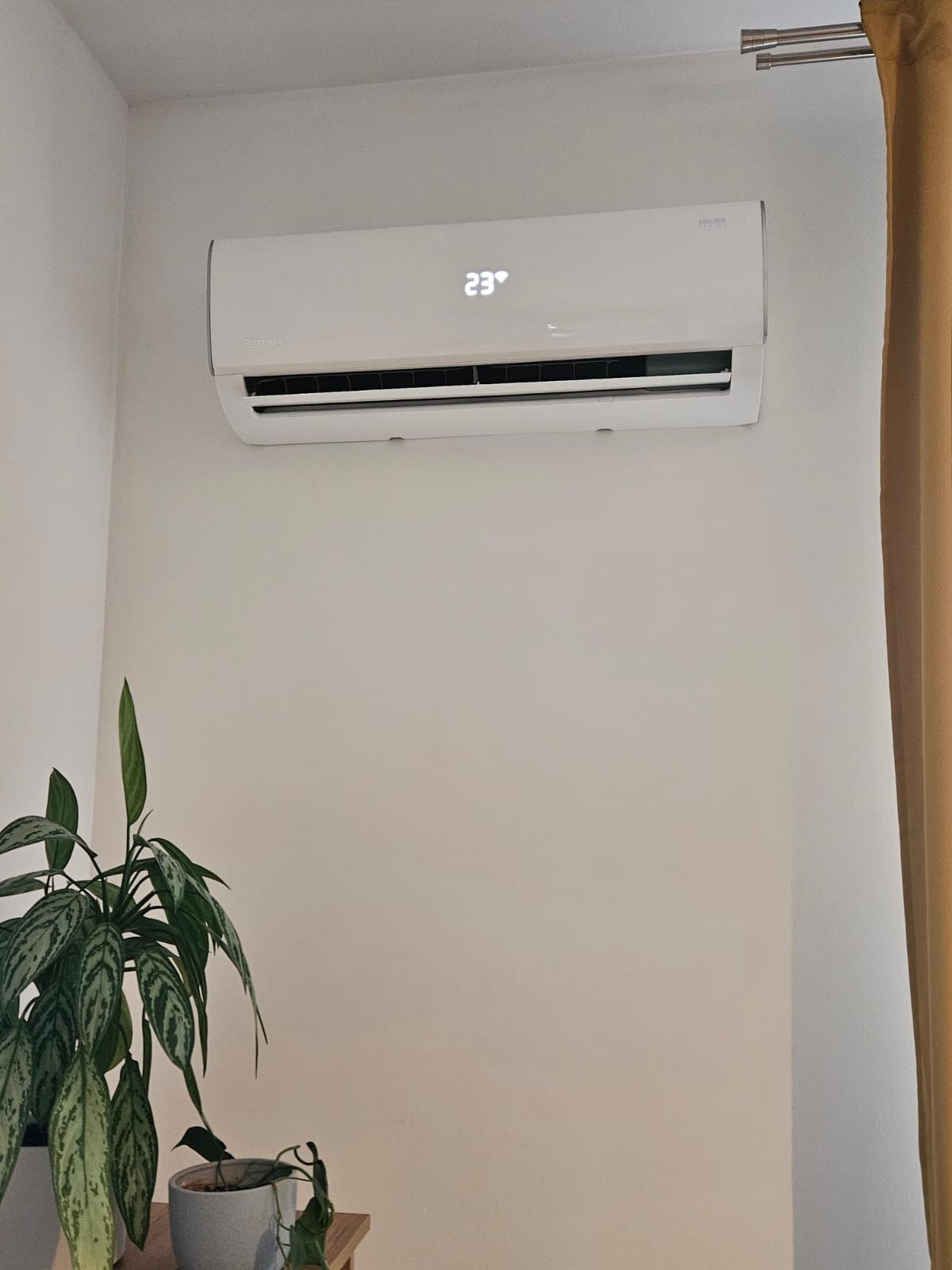 Biały klimatyzator Rotenso z wyświetlaną temperaturą 23°C na ścianie w jasnym pomieszczeniu, obok zasłony i rośliny doniczkowej.