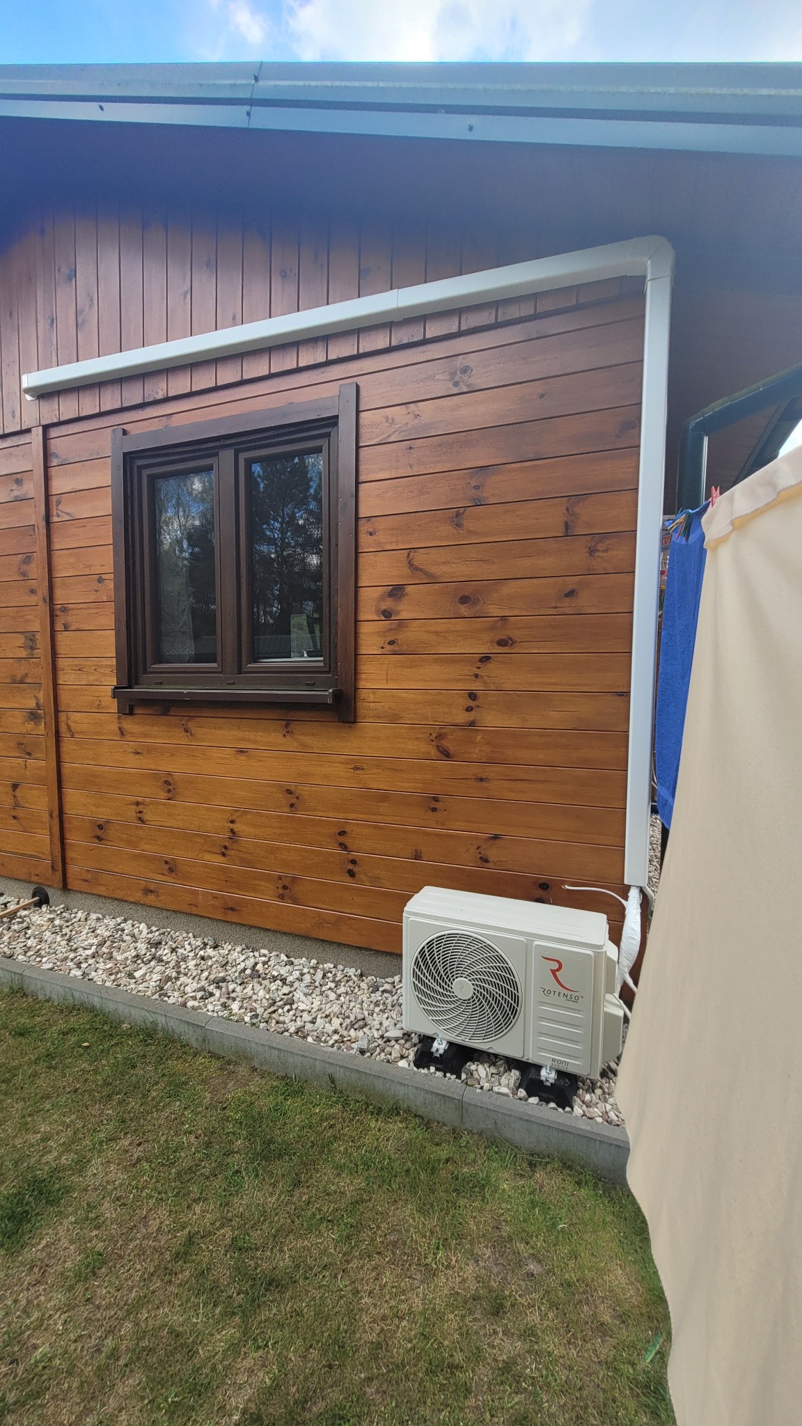 Biała jednostka zewnętrzna klimatyzacji Rotenso, zamontowana przy drewnianym domku z brązowymi oknami i białymi obramowaniami, na tle zielonej trawy i kamieni.