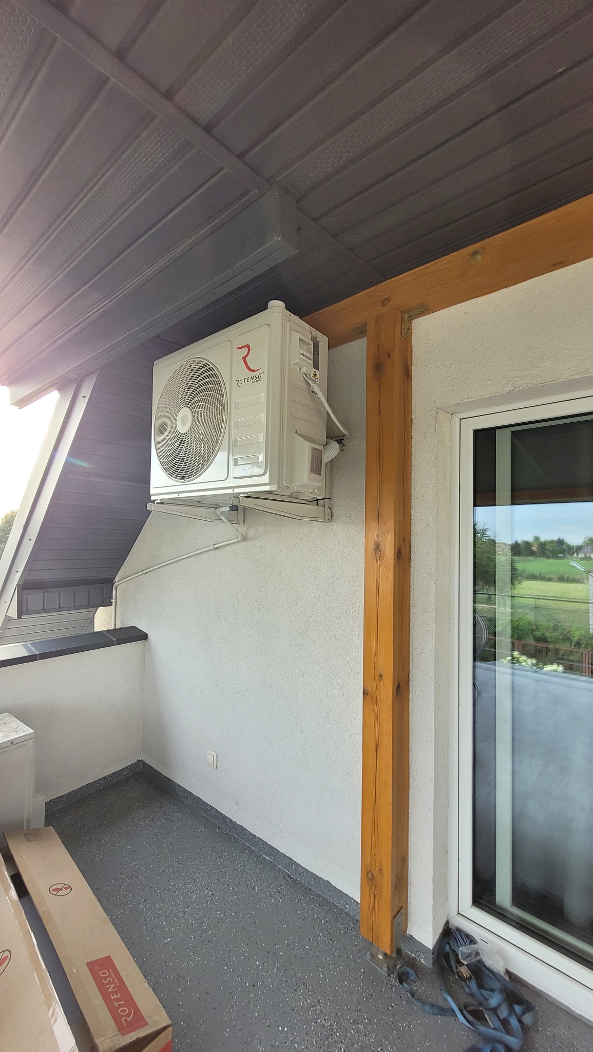 Biała klimatyzacja Rotenso zamontowana na balkonie, widok z dołu, drewniany element konstrukcyjny obok okna balkonowego, szare podłoże balkonu.