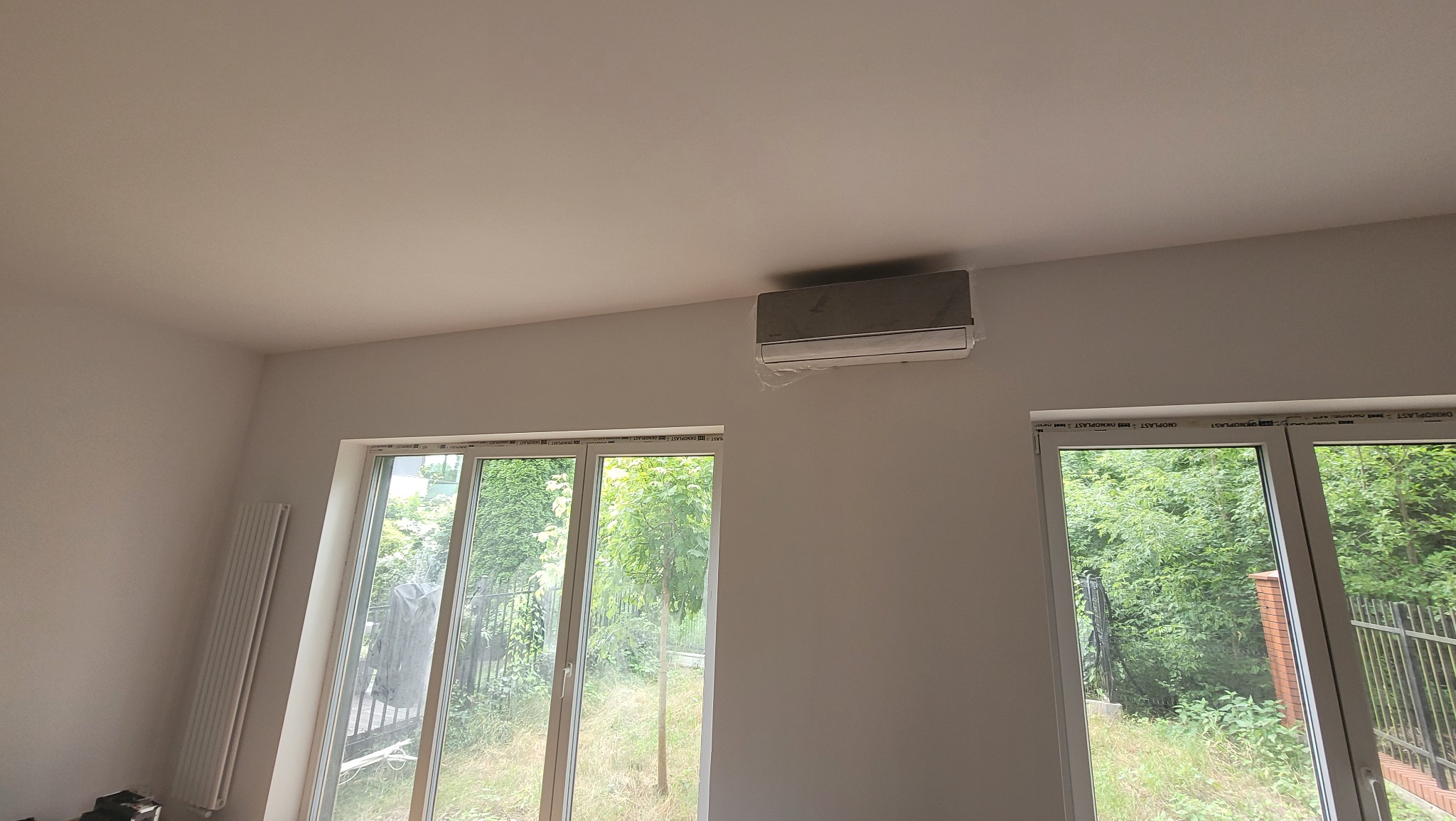 Nowoczesny klimatyzator na ścianie w jasnym pokoju z dużymi oknami i widokiem na zieleń. Klimatyzacja w folii ochronnej po montażu. Minimalistyczny design wnętrza.