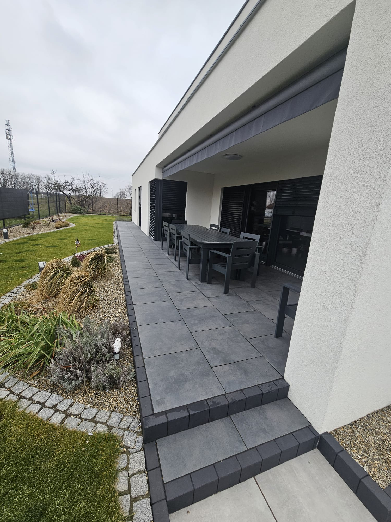 Nowoczesny taras z szarych płyt, obrzeża z kostki granitowej, meble ogrodowe w odcieniach szarości. Widok na zadbany ogród z elementami dekoracyjnymi i trawami ozdobnymi.