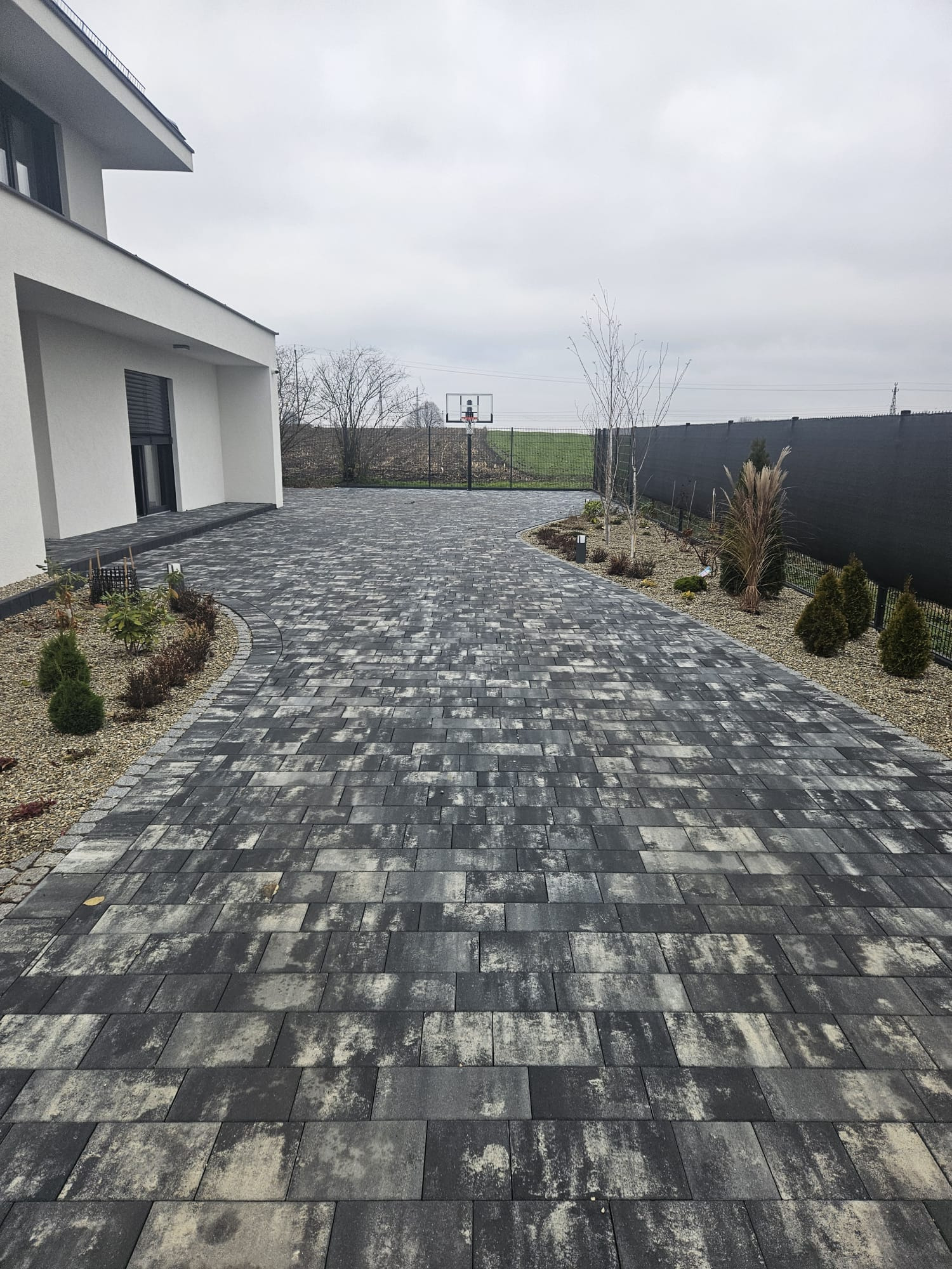 Ułożona kostka brukowa w odcieniach szarości prowadząca do domu z elementami zieleni i koszem do koszykówki w tle, tworząca estetyczny podjazd.