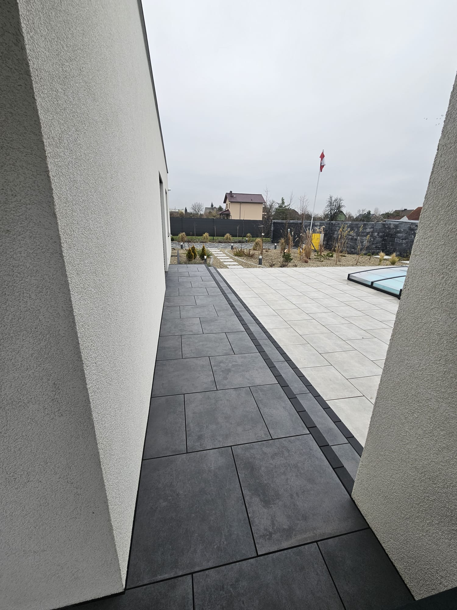 Nowoczesna aranżacja: ciemnoszara kostka brukowa prowadzi do jasnego patio z przykryciem basenu. Minimalistyczny design, neutralna kolorystyka, flaga w tle.