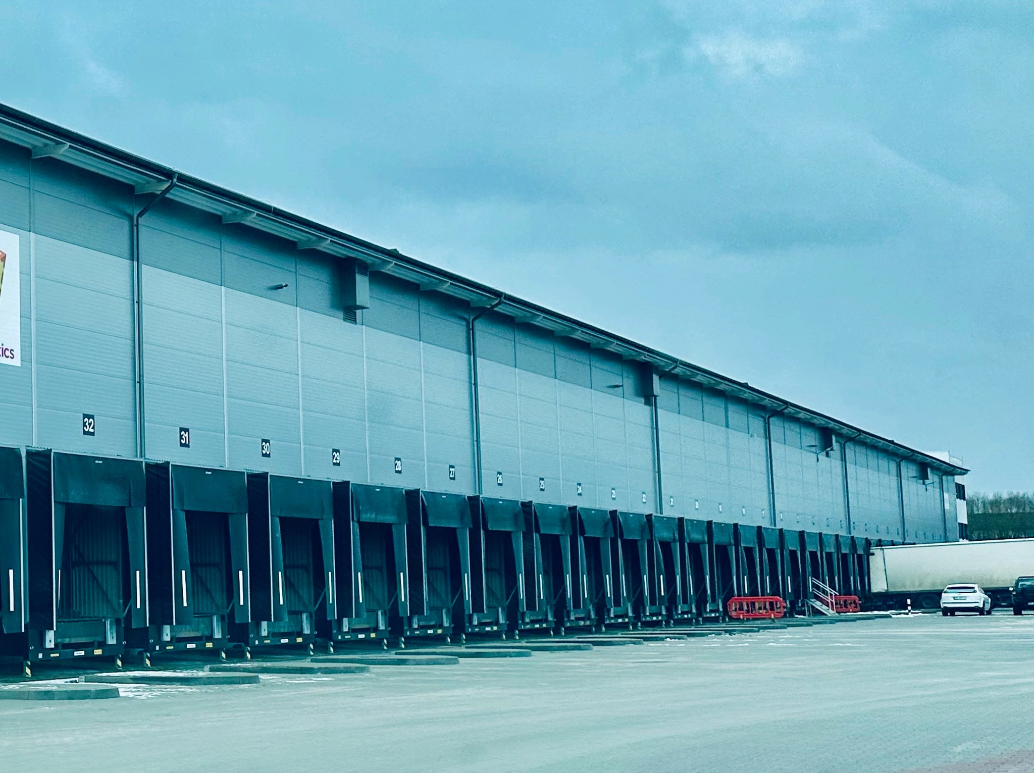 Nowoczesna hala magazynowa z rampami załadunkowymi, widok z zewnątrz. Na parkingu zaparkowane samochody. Szara elewacja budynku i błękitne niebo w tle.