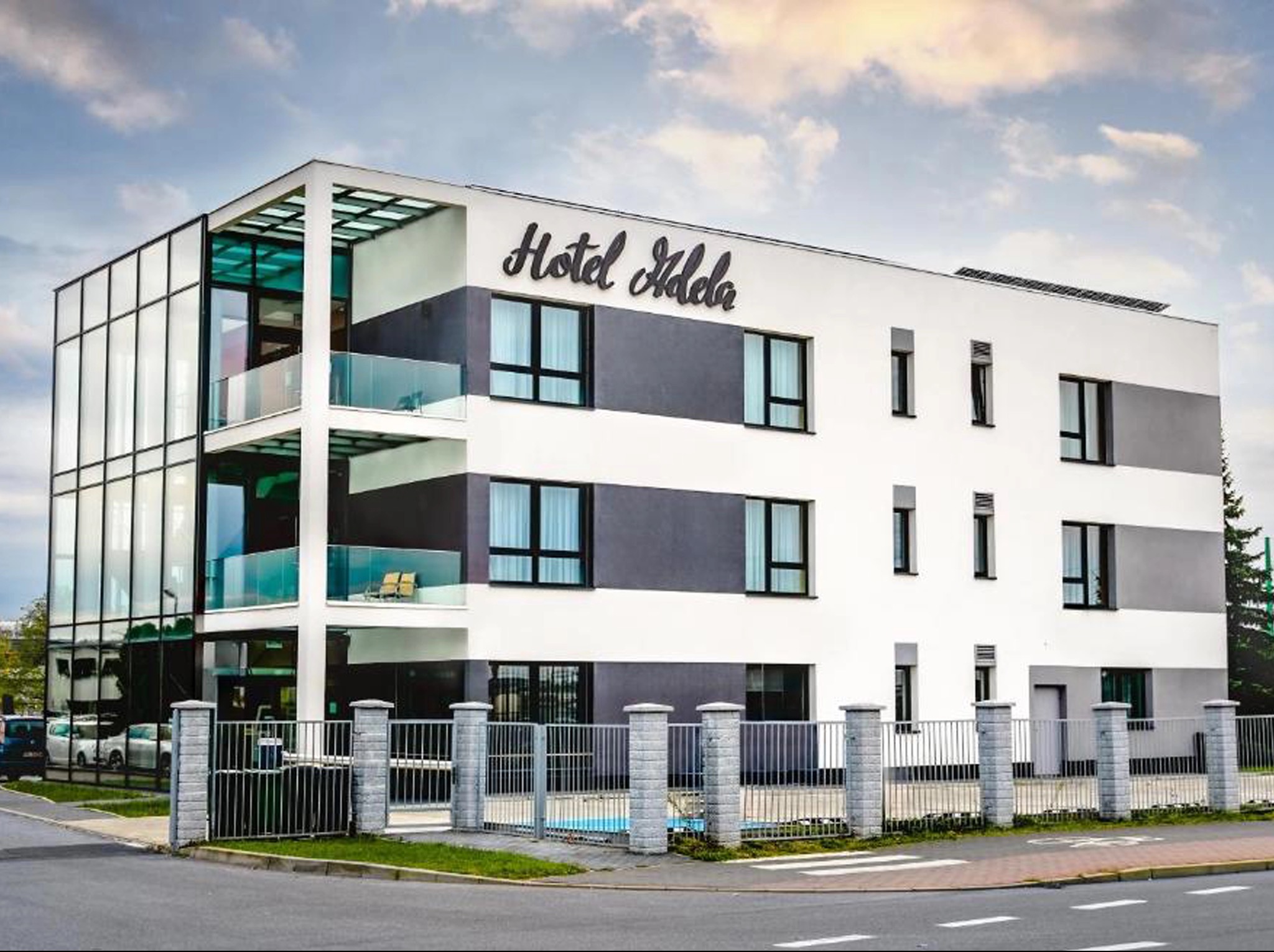 Nowoczesny budynek hotelu Adela z przeszkloną klatką schodową, minimalistyczną bryłą i szklanymi tarasami. Widoczna elewacja w odcieniach bieli i szarości.