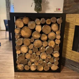 Metalowa rama wypełniona drewnem kominkowym, tworząca dekoracyjny element wnętrza. U góry wazon z lawendą. Obok widoczny fragment obudowy kominka.